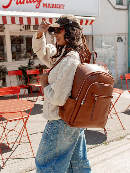 Woman with a brown vegan leather backpack walking past a market with red and white striped awning.